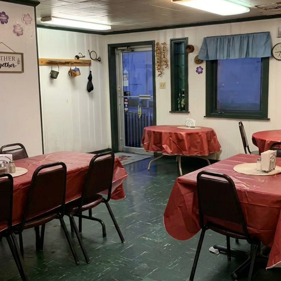 Cozy restaurant dining room with red tablecloths, black chairs, floral decorations, curtained windows, and a Gather Together sign.