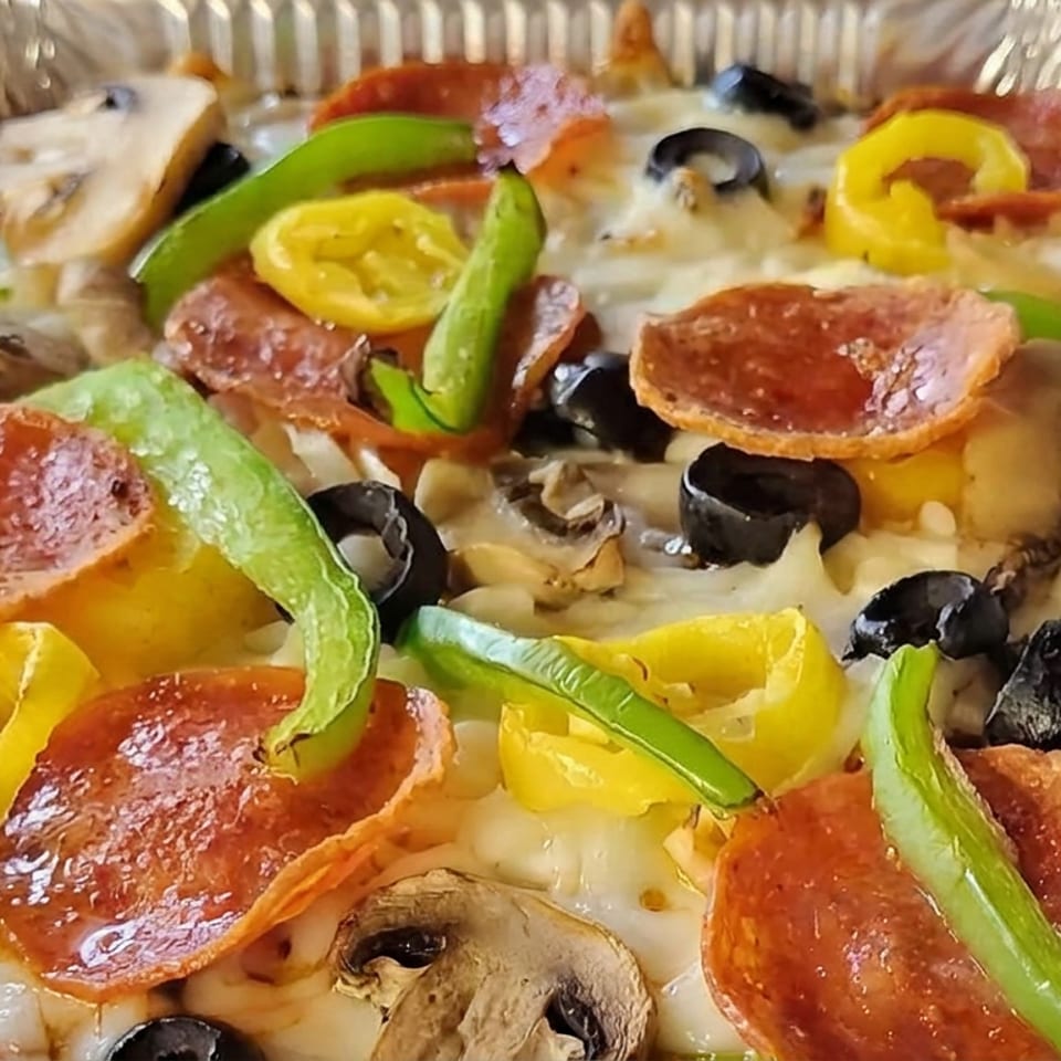 Close-up of a pepperoni pizza topped with melted cheese, green and yellow peppers, black olives, mushrooms, and onions in a foil tray.