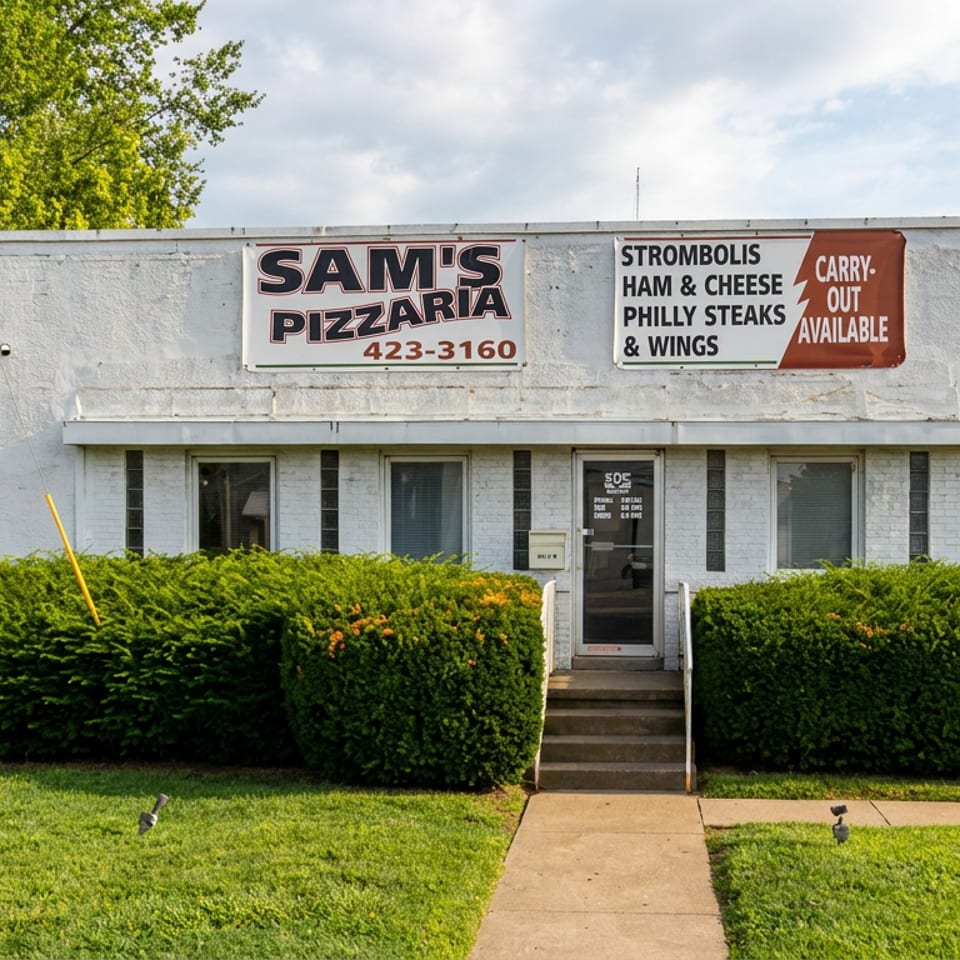 Street view of Sam’s Pizzaria, a small white building with trimmed hedges, outdoor signage, and a central entrance.
