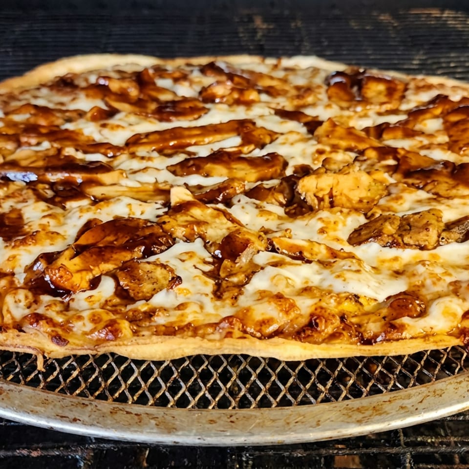 Thin-crust pizza topped with melted cheese, grilled chicken, and glossy brown sauce on a mesh baking tray.