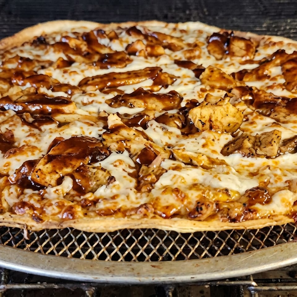 Thin-crust pizza topped with melted cheese, grilled chicken pieces, and glossy barbecue sauce on a metal pizza screen.