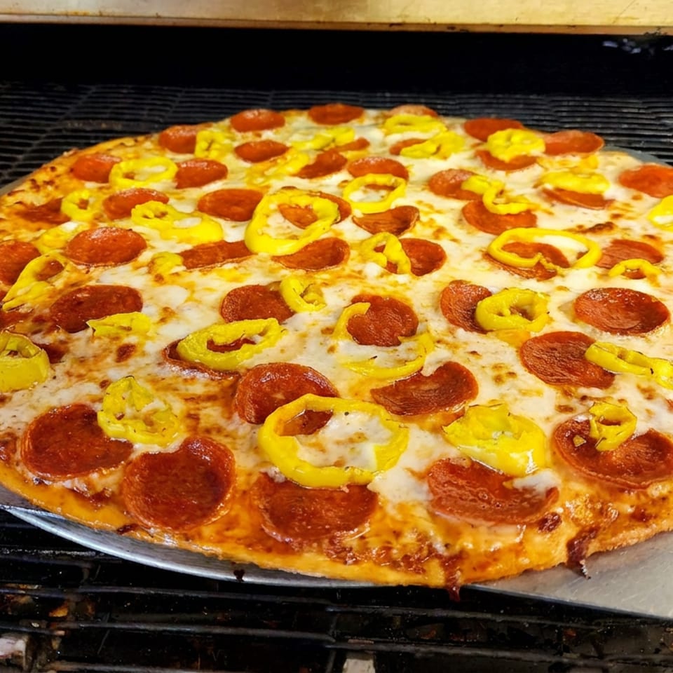 Large freshly baked pepperoni pizza topped with melted cheese and yellow peppers, resting on a metal peel inside a pizza oven.